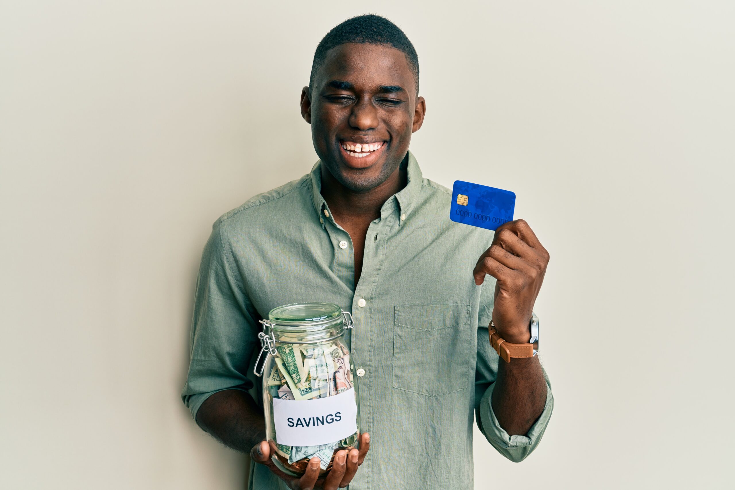 young african american man holding credit card and jar with dollars smiling and laughing hard out loud because funny crazy joke.